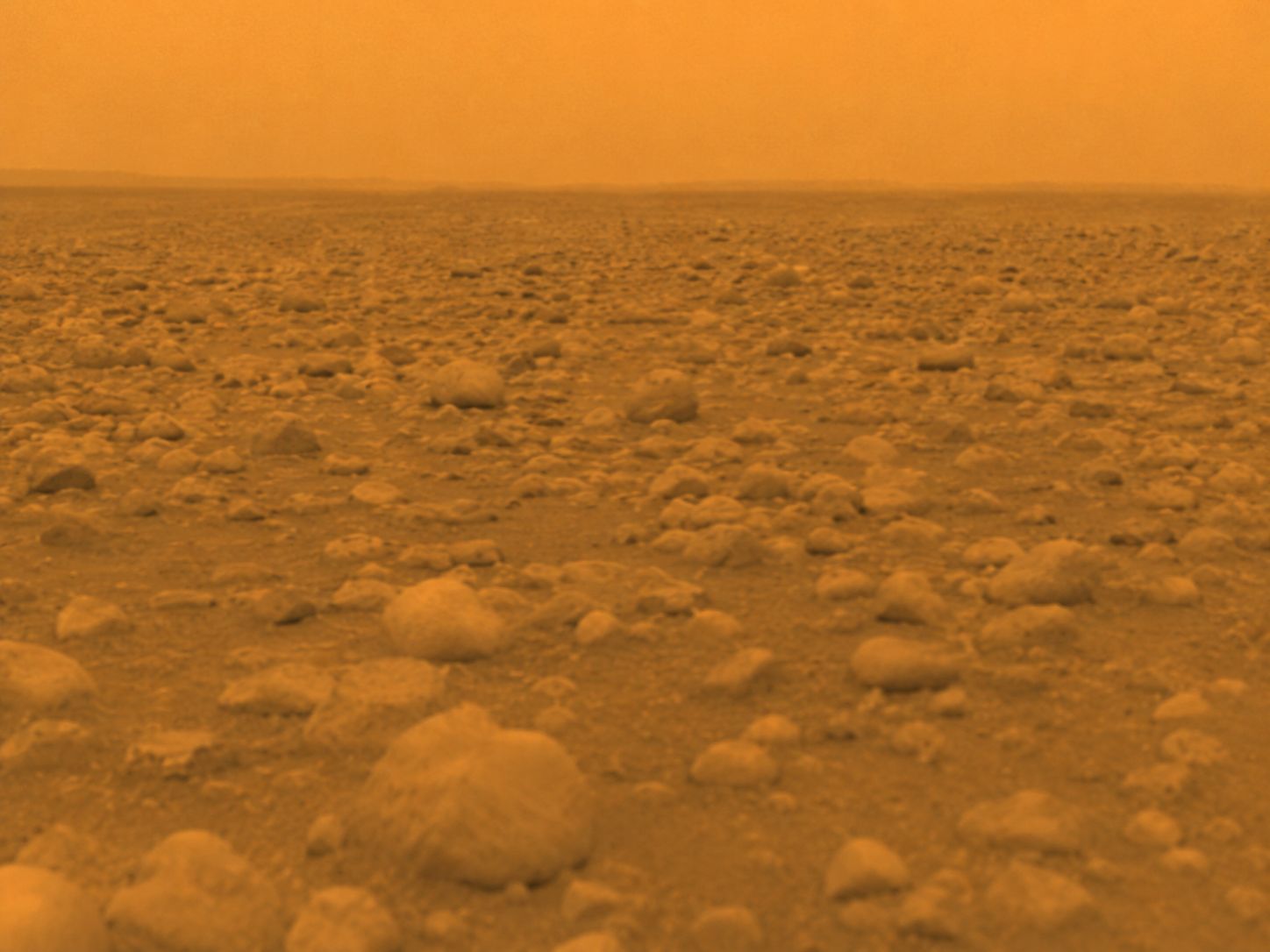 Photograph from the Huygens probe of Titan's surface, showing pale rounded rocks of water ice scattered across an orange plain